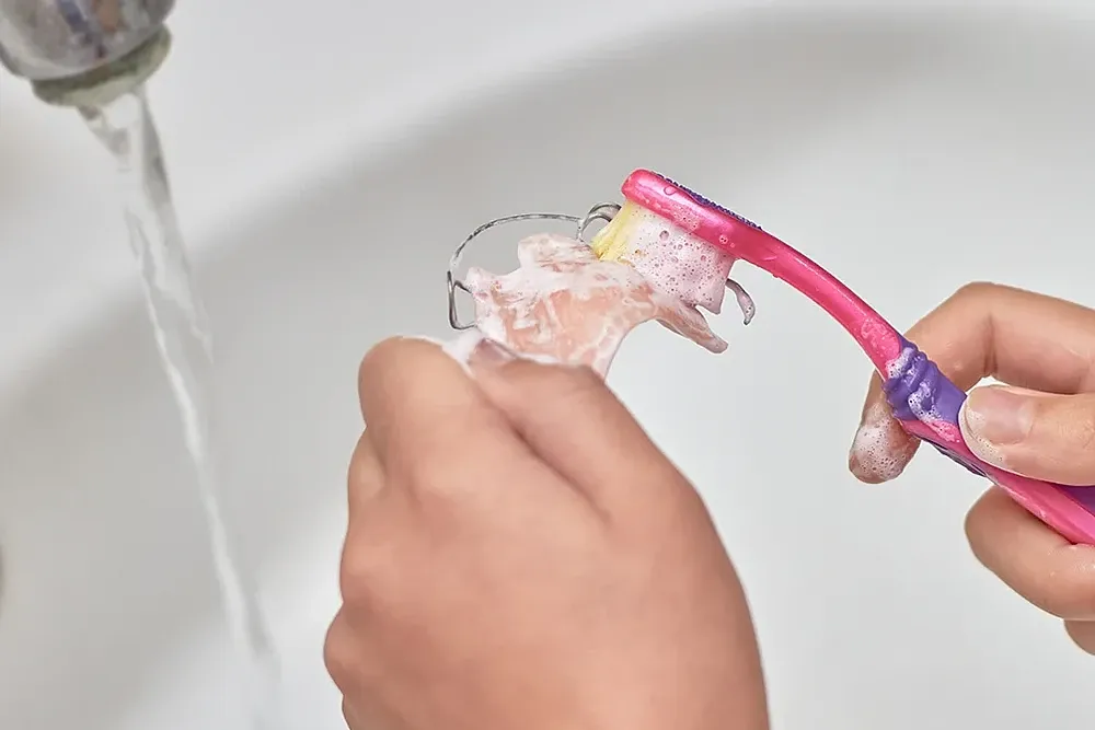 Kind reinigt eine herausnehmbare Zahnspange mit einer Zahnbürste unter fließendem Wasser.