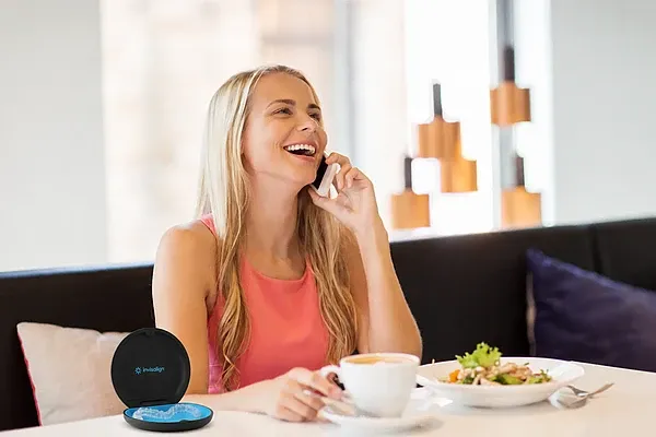 Lächelnde Frau telefoniert in einem Café; auf dem Tisch stehen Kaffee, Salat und eine geöffnete Invisalign-Box mit Alignern.